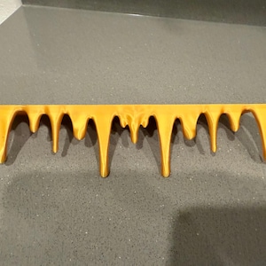 Peut inclure: Une pi&egrave;ce d&eacute;corative horizontale jaune dor&eacute; avec un motif d&eacute;goulinant. La pi&egrave;ce a un bord sup&eacute;rieur plat et de nombreuses gouttes arrondies descendantes de longueurs variables. L'objet est fait d'un mat&eacute;riau solide et est pos&eacute; sur un fond gris.