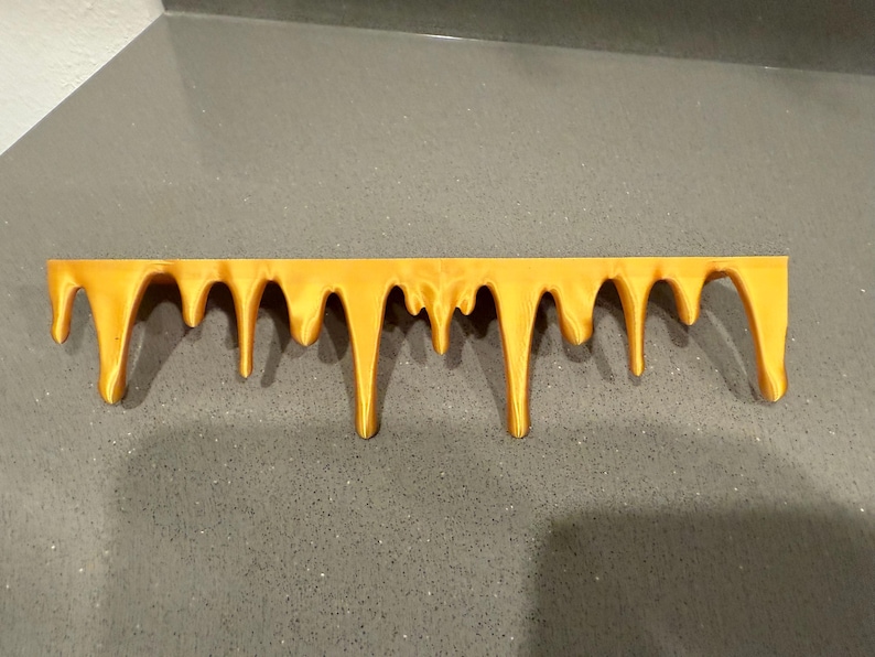 Peut inclure: Une pi&egrave;ce d&eacute;corative horizontale jaune dor&eacute; avec un motif d&eacute;goulinant. La pi&egrave;ce a un bord sup&eacute;rieur plat et de nombreuses gouttes arrondies descendantes de longueurs variables. L'objet est fait d'un mat&eacute;riau solide et est pos&eacute; sur un fond gris.