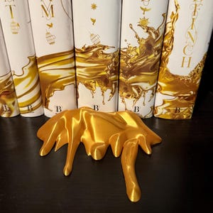 May include: A golden-yellow, abstract sculpture with a flowing, liquid-like design. The sculpture is in front of a row of books with white spines and gold accents. The books have the letter "B" on the bottom.