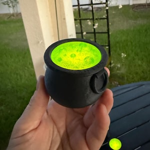 May include: A black cauldron with a green glowing liquid inside. The cauldron is made of plastic and has a handle on the side.