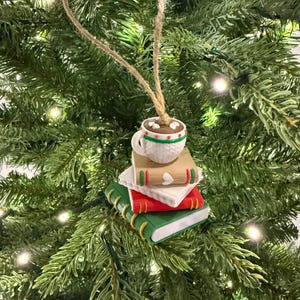 K&ouml;nnte beinhalten: Ein Weihnachtsbaumschmuck mit einem Stapel aus drei Miniaturb&uuml;chern, gr&uuml;n, rot und beige, gekr&ouml;nt von einer Tasse hei&szlig;er Schokolade. Die Tasse ist wei&szlig; mit gr&uuml;nen und roten Verzierungen. Der Schmuck h&auml;ngt an einer Schnur.