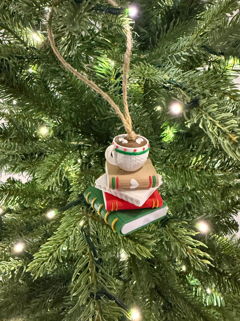 K&ouml;nnte beinhalten: Ein Weihnachtsbaumschmuck mit einem Stapel aus drei Miniaturb&uuml;chern, gr&uuml;n, rot und beige, gekr&ouml;nt von einer Tasse hei&szlig;er Schokolade. Die Tasse ist wei&szlig; mit gr&uuml;nen und roten Verzierungen. Der Schmuck h&auml;ngt an einer Schnur.