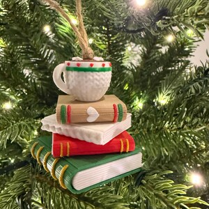 K&ouml;nnte beinhalten: Ein Weihnachtsbaumschmuck mit einem Stapel Miniaturb&uuml;cher, gekr&ouml;nt von einer Kaffeetasse. Die B&uuml;cher sind rot, gr&uuml;n und beige mit goldfarbenen Buchr&uuml;cken. Die Kaffeetasse ist wei&szlig; mit gr&uuml;n-rotem Rand.