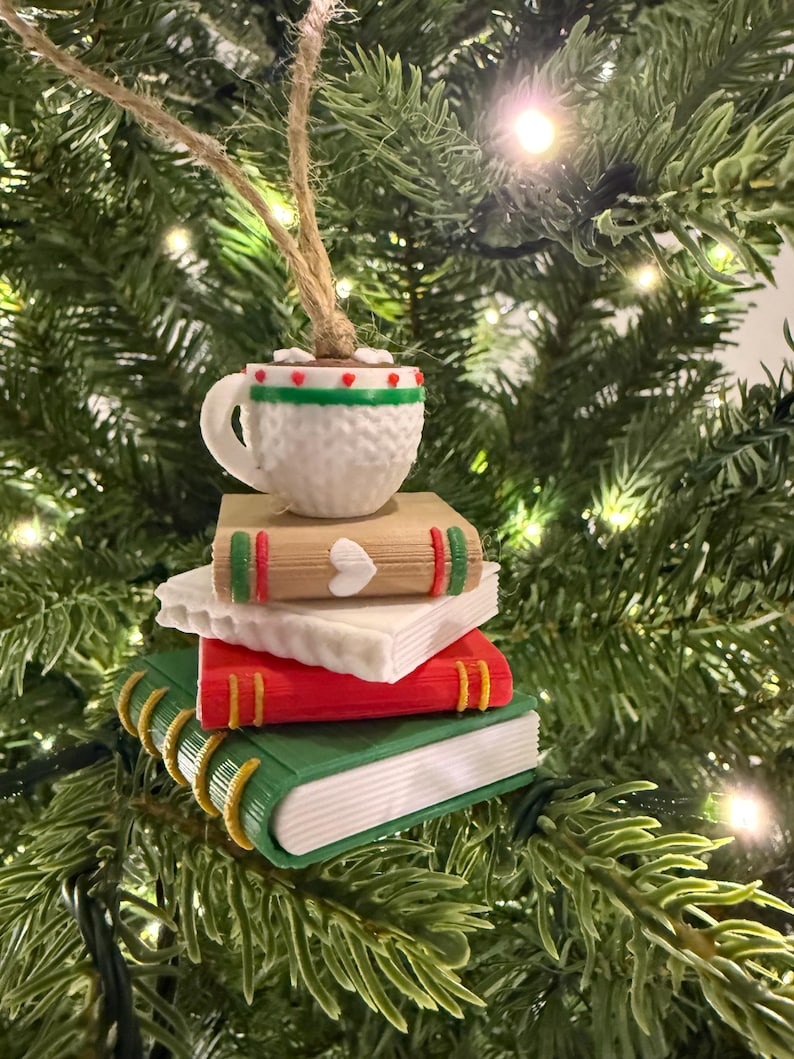 K&ouml;nnte beinhalten: Ein Weihnachtsbaumschmuck mit einem Stapel Miniaturb&uuml;cher, gekr&ouml;nt von einer Kaffeetasse. Die B&uuml;cher sind rot, gr&uuml;n und beige mit goldfarbenen Buchr&uuml;cken. Die Kaffeetasse ist wei&szlig; mit gr&uuml;n-rotem Rand.