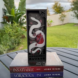 May include: A black rectangular decorative piece featuring a white snake and red heart design, set atop three books. The books are titled "Insatiable", "Voracious", and "Restitution" by Leigh Rivers. The piece is likely a bookend or decorative item.