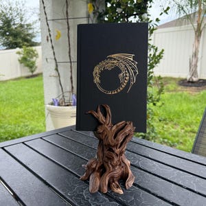 Könnte beinhalten: Ein schwarzes Hardcover-Buch mit einem goldfarbenen, kreisförmigen Drachenemblem auf dem Einband, das in einem braunen, strukturierten Ständer in Form eines Baumstumpfes steht, der mit tiefen Rillen und organischen Formen an knorriges Holz erinnert.