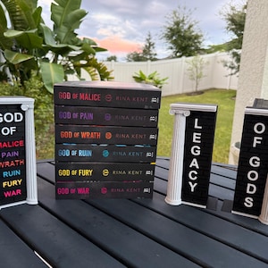 May include: A stack of six books with black covers, titled "God of Malice," "God of Pain," "God of Wrath," "God of Ruin," "God of Fury," and "God of War." Decorative letter boards spell out "God of Malice," "Legacy," and "Of Gods."