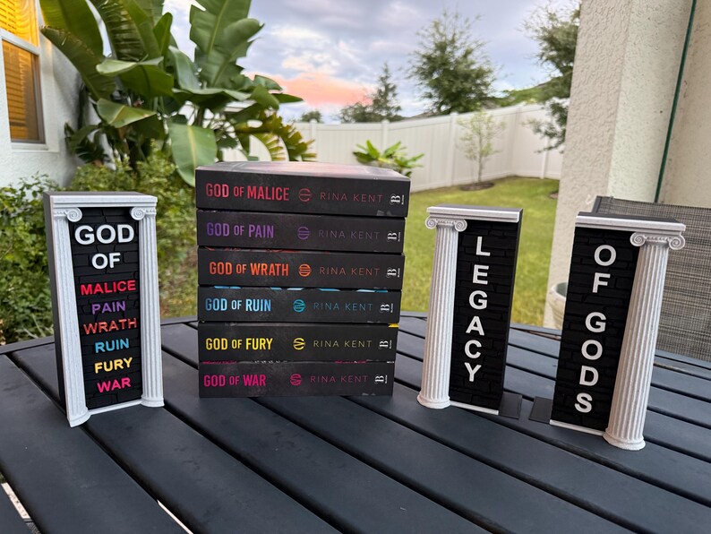 May include: A stack of six books with black covers, titled "God of Malice," "God of Pain," "God of Wrath," "God of Ruin," "God of Fury," and "God of War." Decorative letter boards spell out "God of Malice," "Legacy," and "Of Gods."