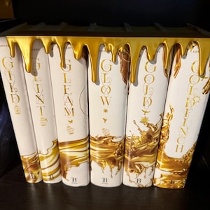 Peut inclure: Une collection de livres &agrave; couverture rigide avec des couvertures blanches et des lettres dor&eacute;es. Les titres incluent "GILD", "GLINT", "GLEAM", "GLOW" et "GOLDENCH". Un &eacute;l&eacute;ment d&eacute;coratif dor&eacute; d&eacute;gouline sur le dessus des livres.