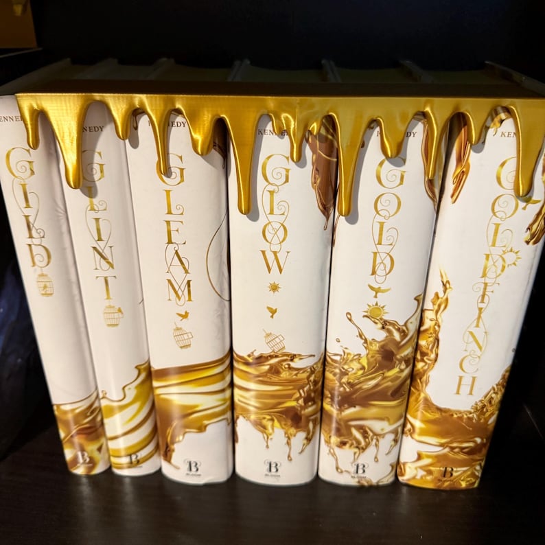 Peut inclure: Une collection de livres &agrave; couverture rigide avec des couvertures blanches et des lettres dor&eacute;es. Les titres incluent "GILD", "GLINT", "GLEAM", "GLOW" et "GOLDENCH". Un &eacute;l&eacute;ment d&eacute;coratif dor&eacute; d&eacute;gouline sur le dessus des livres.