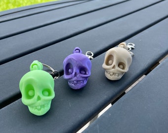 May include: Three small skull keychains in different colors: green, purple, and beige. The keychains are on a dark gray wooden surface.
