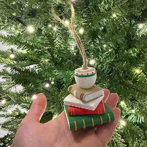 K&ouml;nnte beinhalten: Ein Weihnachtsbaumschmuck mit einem Stapel Miniaturb&uuml;cher in Rot, Wei&szlig; und Gr&uuml;n, gekr&ouml;nt von einem kleinen wei&szlig;en Topf mit einem gr&uuml;nen und roten Muster. Der Schmuck wird von einer Schnurschlaufe gehalten.