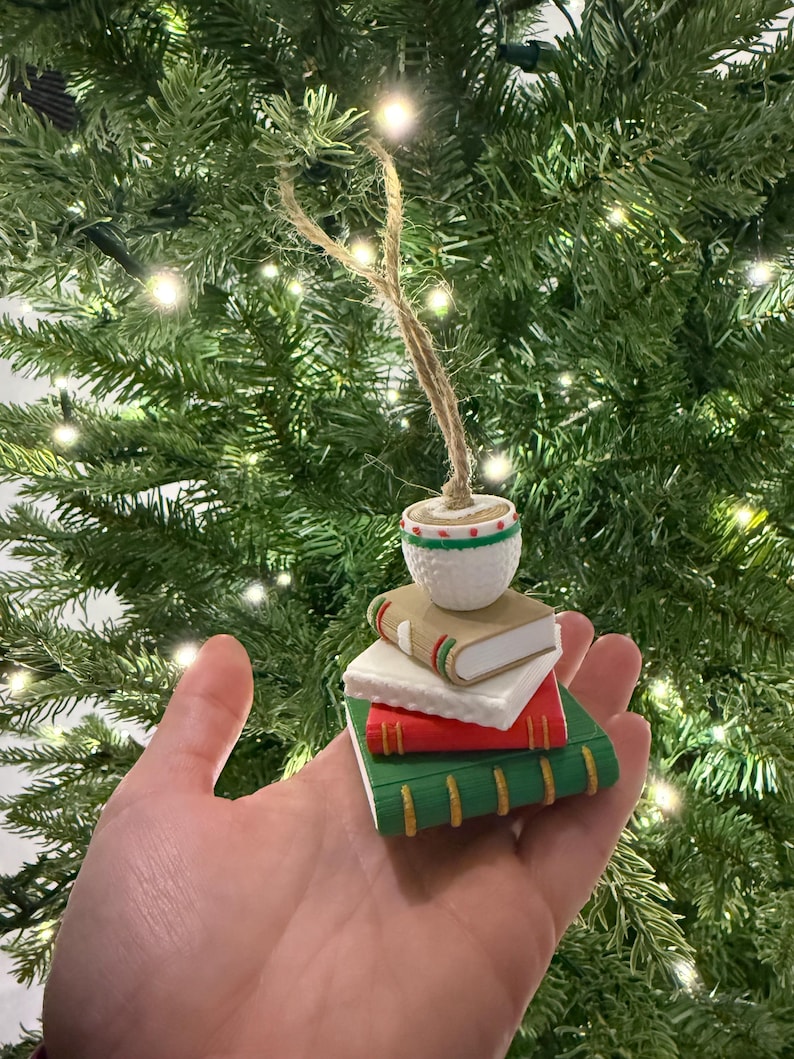 K&ouml;nnte beinhalten: Ein Weihnachtsbaumschmuck mit einem Stapel Miniaturb&uuml;cher in Rot, Wei&szlig; und Gr&uuml;n, gekr&ouml;nt von einem kleinen wei&szlig;en Topf mit einem gr&uuml;nen und roten Muster. Der Schmuck wird von einer Schnurschlaufe gehalten.