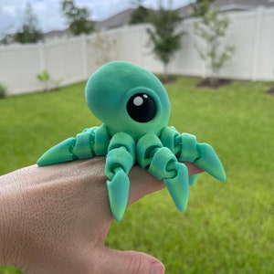 May include: A green and blue octopus figurine with a single large black eye. The octopus is posed with its tentacles outstretched, as if it is crawling.