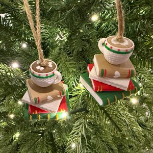 K&ouml;nnte beinhalten: Weihnachtsbaumschmuck mit einer wei&szlig;en Tasse mit braunem Getr&auml;nk und Marshmallows auf einem Stapel B&uuml;cher. Die B&uuml;cher haben rote, gr&uuml;ne und beige Einb&auml;nde. Die Ornamente h&auml;ngen an Schn&uuml;ren vor einem gr&uuml;nen Weihnachtsbaum.