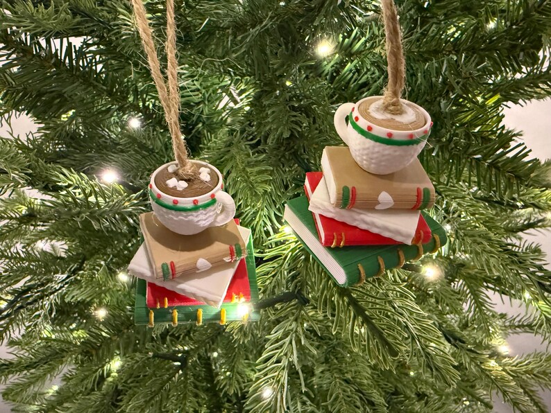 K&ouml;nnte beinhalten: Weihnachtsbaumschmuck mit einer wei&szlig;en Tasse mit braunem Getr&auml;nk und Marshmallows auf einem Stapel B&uuml;cher. Die B&uuml;cher haben rote, gr&uuml;ne und beige Einb&auml;nde. Die Ornamente h&auml;ngen an Schn&uuml;ren vor einem gr&uuml;nen Weihnachtsbaum.