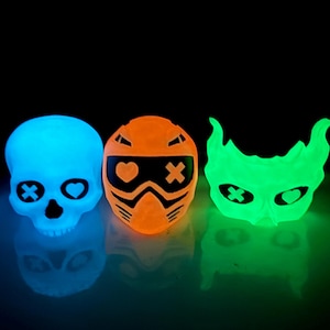 May include: Three glowing decorative objects: a blue skull with heart and cross details, an orange helmet-shaped design with a heart and cross, and a green mask-like shape with similar details. All have a reflective surface.