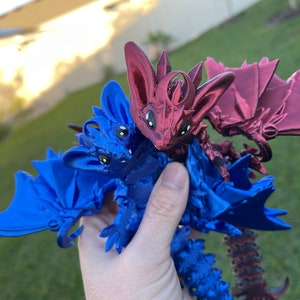 May include: A pair of 3D-printed fantasy creatures, one blue and one maroon, with bat-like wings and large eyes. The creatures are held in a hand against a blurred green background.
