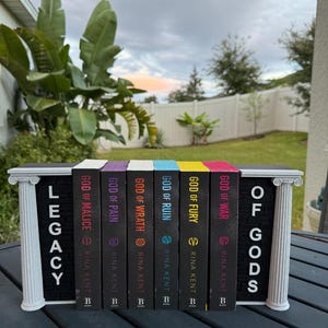 May include: A collection of seven books titled "Legacy of Gods" by Rina Kent, displayed in a decorative white and black bookend. The book spines are black with colorful accents and titles like "God of War" and "God of Fury".