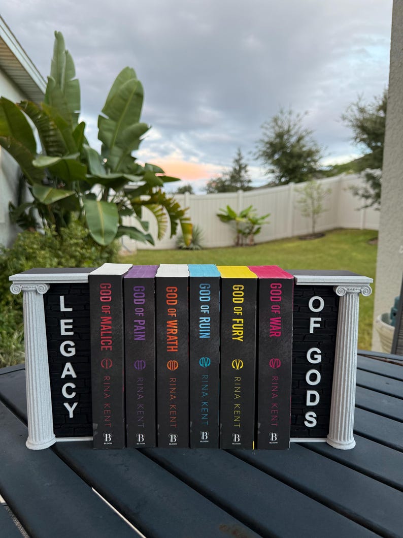 May include: A collection of seven books titled "Legacy of Gods" by Rina Kent, displayed in a decorative white and black bookend. The book spines are black with colorful accents and titles like "God of War" and "God of Fury".
