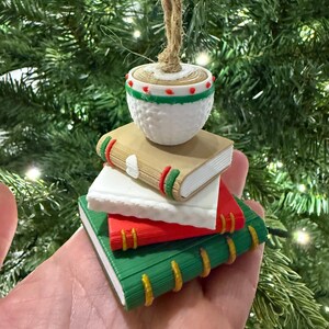 K&ouml;nnte beinhalten: Ein Weihnachtsornament mit einem Stapel Miniaturb&uuml;cher in Gr&uuml;n, Rot und Wei&szlig;, gekr&ouml;nt von einer wei&szlig;en, strukturierten Kugel. Die B&uuml;cher haben farbige Buchr&uuml;cken und das Ornament h&auml;ngt an einer nat&uuml;rlichen Schnurschlaufe, ideal f&uuml;r die Weihnachtsdekoration.
