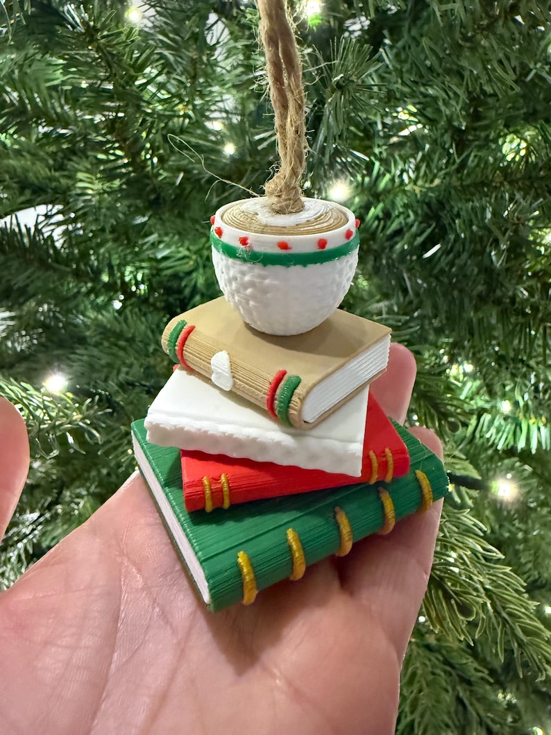 K&ouml;nnte beinhalten: Ein Weihnachtsornament mit einem Stapel Miniaturb&uuml;cher in Gr&uuml;n, Rot und Wei&szlig;, gekr&ouml;nt von einer wei&szlig;en, strukturierten Kugel. Die B&uuml;cher haben farbige Buchr&uuml;cken und das Ornament h&auml;ngt an einer nat&uuml;rlichen Schnurschlaufe, ideal f&uuml;r die Weihnachtsdekoration.