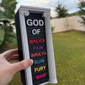 May include: A rectangular black sign with white Greek columns and the words "GOD OF MALICE PAIN WRATH RUIN FURY WAR" in colorful 3D lettering. The sign is held in front of a green lawn and blue sky.