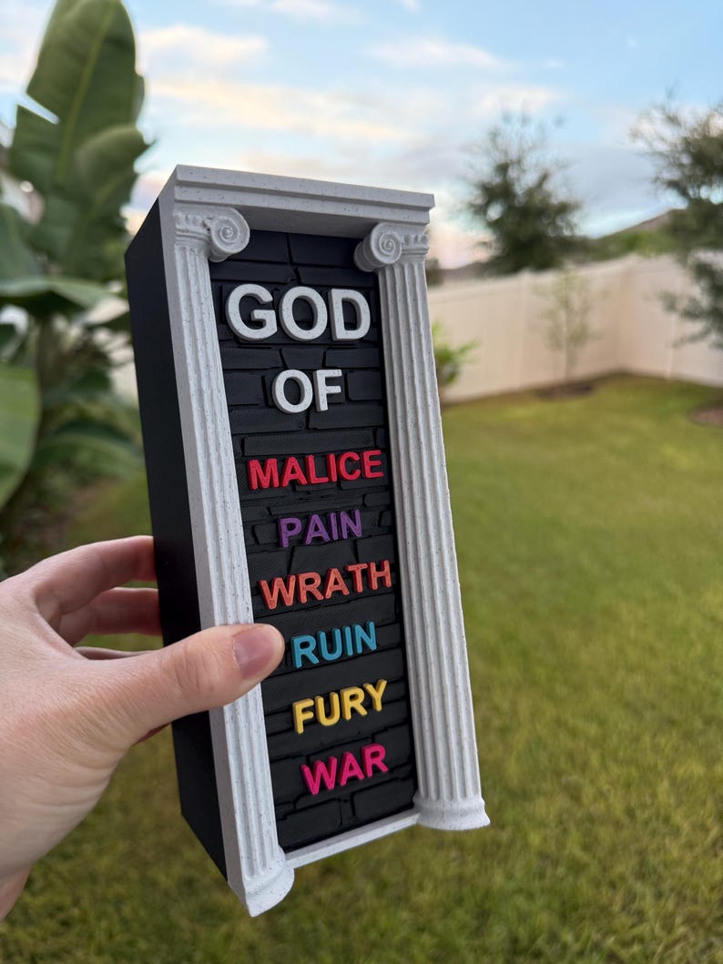May include: A rectangular black sign with white Greek columns and the words "GOD OF MALICE PAIN WRATH RUIN FURY WAR" in colorful 3D lettering. The sign is held in front of a green lawn and blue sky.