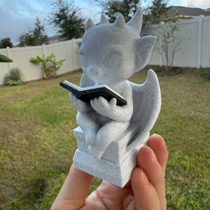 May include: A small, speckled, gray statue of a winged creature reading a book. The statue has horns and is seated on a pedestal. The book is black. The statue is held in a hand against a background of grass, trees, and a white fence.