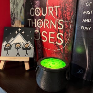 May include: A black cauldron with a green glowing liquid inside. The cauldron is sitting on a wooden surface in front of two books. The book on the left has the title "A Court of Thorns and Roses" and the book on the right has the title "A Court of Mist and Fury".