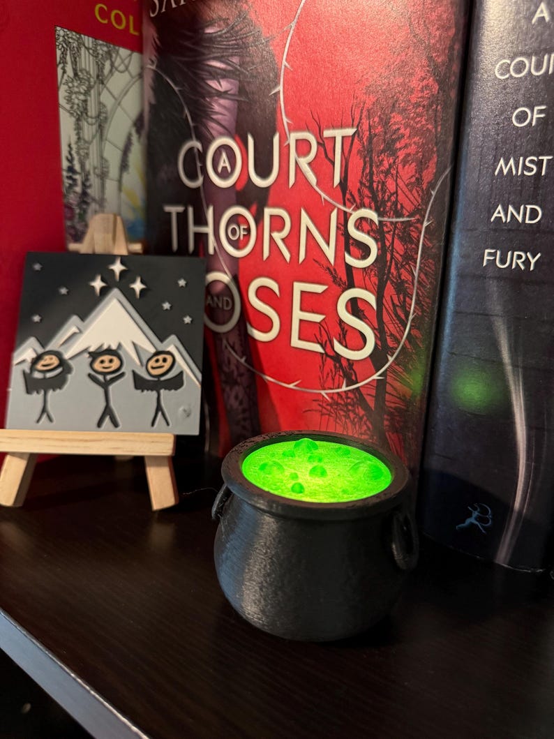 May include: A black cauldron with a green glowing liquid inside. The cauldron is sitting on a wooden surface in front of two books. The book on the left has the title "A Court of Thorns and Roses" and the book on the right has the title "A Court of Mist and Fury".