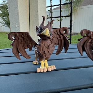 May include: A brown griffin figurine with outstretched wings, yellow feet, and a yellow beak. The griffin has a detailed, textured appearance and is positioned on a dark blue surface. The background includes a trellis and a glimpse of a house.