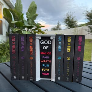 May include: A collection of hardcover books with black spines, featuring titles like "God of Malice" and "God of War." The books are arranged on a dark surface, with a decorative white structure in the center. The author's name, Rina Kent, is printed on each book.