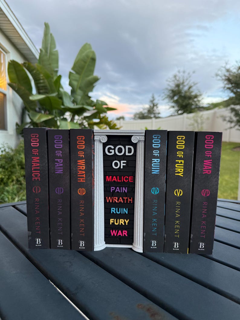 May include: A collection of hardcover books with black spines, featuring titles like "God of Malice" and "God of War." The books are arranged on a dark surface, with a decorative white structure in the center. The author's name, Rina Kent, is printed on each book.