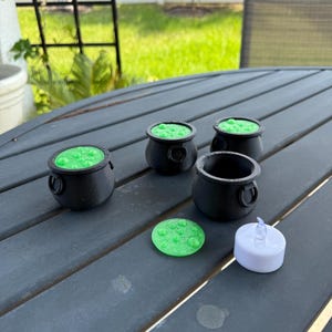 May include: Four black cauldron-shaped candle holders with green wax and a green wax disc and a white tea light candle on a gray wooden table.