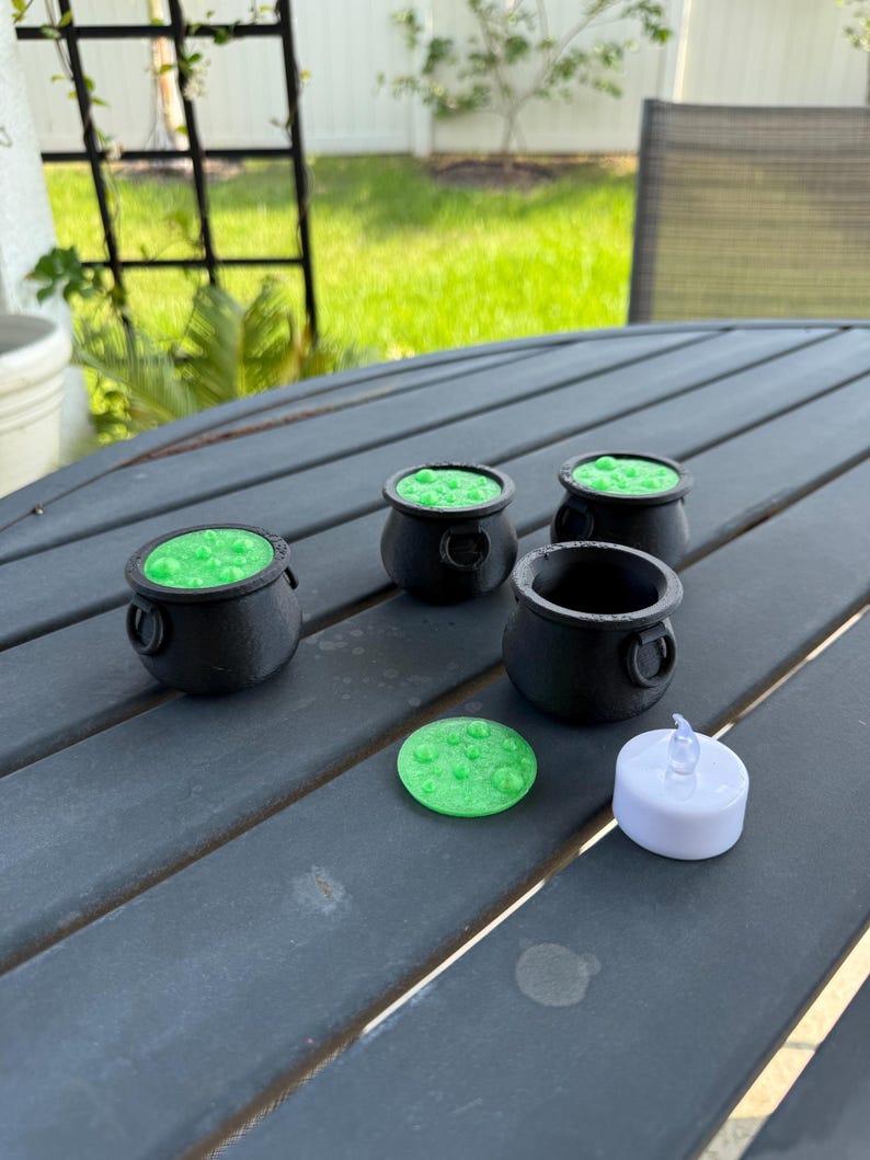 May include: Four black cauldron-shaped candle holders with green wax and a green wax disc and a white tea light candle on a gray wooden table.