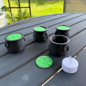 May include: Four black cauldron-shaped candle holders with green wax and a single white tea light candle. The candle holders are on a gray table.