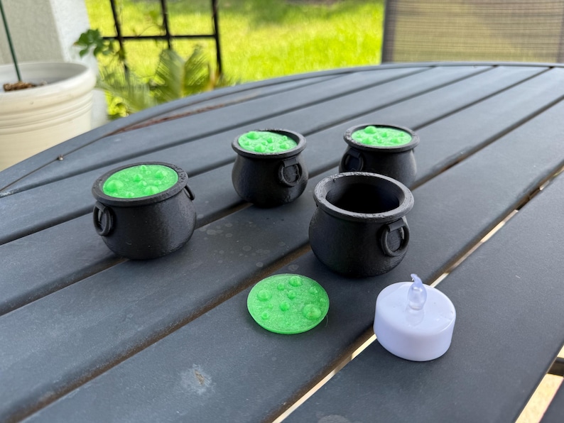 May include: Four black cauldron-shaped candle holders with green wax and a single white tea light candle. The candle holders are on a gray table.
