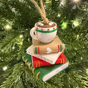 K&ouml;nnte beinhalten: Ein Weihnachtsbaumschmuck mit einer wei&szlig;en Tasse hei&szlig;er Schokolade mit Marshmallows auf einem Stapel von B&uuml;chern. Die B&uuml;cher sind rot, gr&uuml;n und braun mit dekorativen Akzenten. Der Schmuck h&auml;ngt an einer nat&uuml;rlichen Schnur.