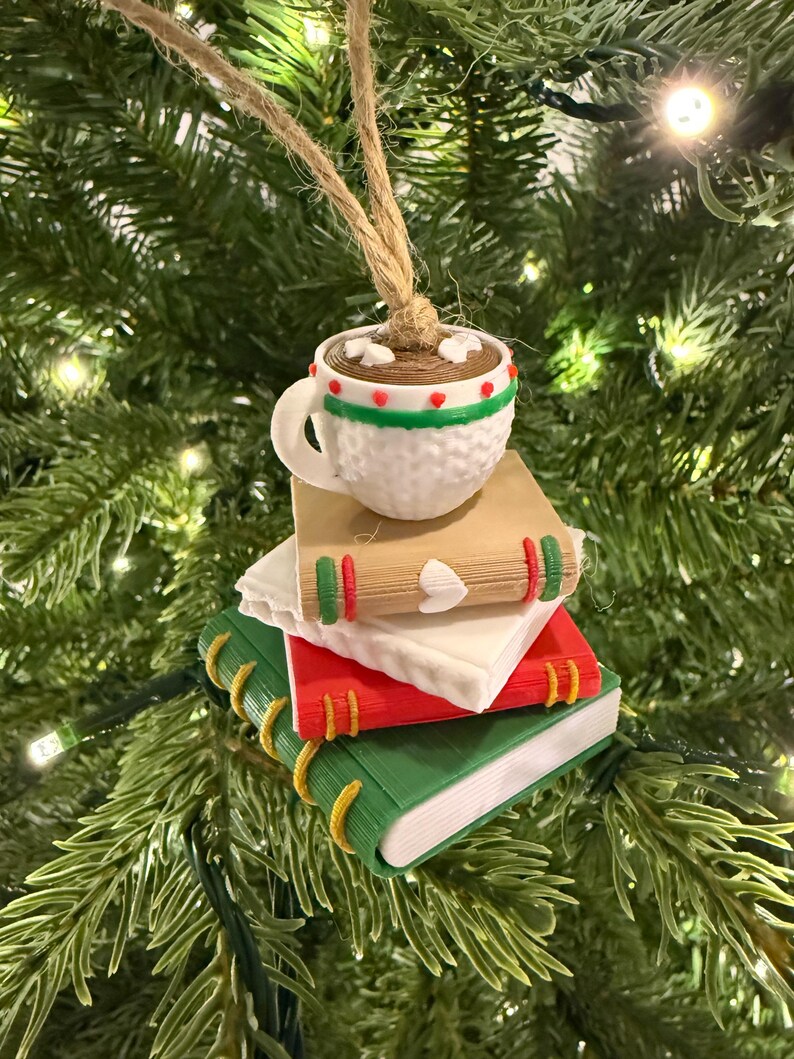 K&ouml;nnte beinhalten: Ein Weihnachtsbaumschmuck mit einer wei&szlig;en Tasse hei&szlig;er Schokolade mit Marshmallows auf einem Stapel von B&uuml;chern. Die B&uuml;cher sind rot, gr&uuml;n und braun mit dekorativen Akzenten. Der Schmuck h&auml;ngt an einer nat&uuml;rlichen Schnur.
