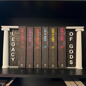 May include: A shelf arrangement featuring a series of books with dark covers. The titles include "God of Malice," "God of Pain," and "God of Wrath." White decorative pillars frame the books, with the words "Legacy" and "Of Gods" displayed.