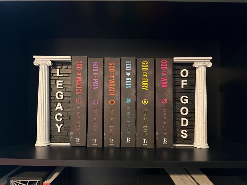 May include: A shelf arrangement featuring a series of books with dark covers. The titles include "God of Malice," "God of Pain," and "God of Wrath." White decorative pillars frame the books, with the words "Legacy" and "Of Gods" displayed.