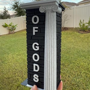 May include: A black and white decorative pillar with the text "OF GODS". The pillar has a textured, brick-like appearance with a white column on the side. The top of the pillar has a decorative design.