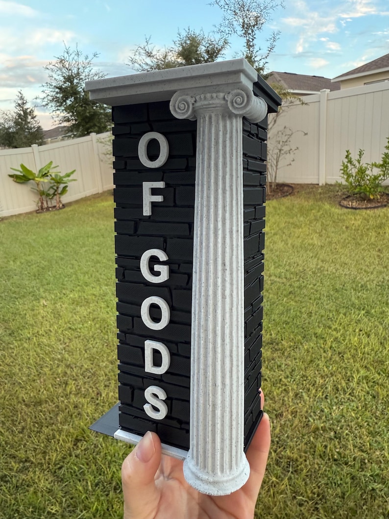 May include: A black and white decorative pillar with the text "OF GODS". The pillar has a textured, brick-like appearance with a white column on the side. The top of the pillar has a decorative design.