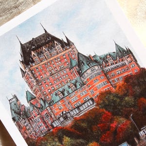May include: Watercolor print of a large, ornate building with a red brick facade, green rooftops, and black turrets. The artwork depicts the building set against a light blue sky, with autumn foliage in the foreground.