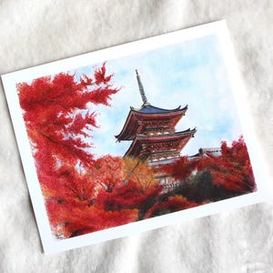 Puede incluir: Pintura a la acuarela de una pagoda japonesa con tejado rojo y azul, rodeada de vibrante follaje oto&ntilde;al rojo. La obra de arte est&aacute; ambientada sobre un cielo azul claro, creando una escena serena y colorida.