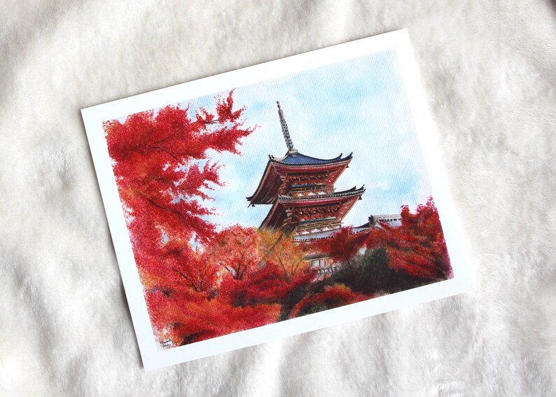Puede incluir: Pintura a la acuarela de una pagoda japonesa con tejado rojo y azul, rodeada de vibrante follaje oto&ntilde;al rojo. La obra de arte est&aacute; ambientada sobre un cielo azul claro, creando una escena serena y colorida.