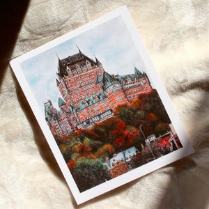 May include: A watercolor print of the Ch&acirc;teau Frontenac in Quebec City. The building is a large, red-brick structure with multiple turrets and green roofs, set against a blue sky and autumn foliage.