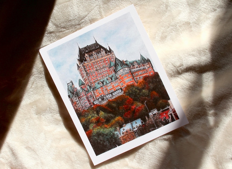 May include: A watercolor print of the Ch&acirc;teau Frontenac in Quebec City. The building is a large, red-brick structure with multiple turrets and green roofs, set against a blue sky and autumn foliage.