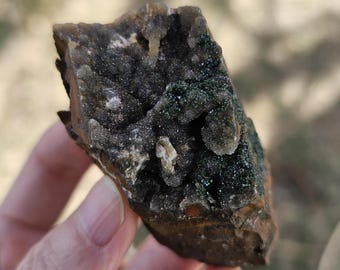 Op de afbeelding: Een ruwe, donkerbruine steen met een cluster van witte en groene kristallen. De kristallen zijn in een natuurlijke formatie geclusterd.